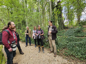 People carrying cameras gather around a man wearing Wildlife Trust branded clothing in a wooded area