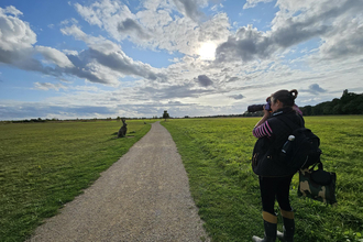 A woman holds her camera up to her face, framing a shot of a dramatic, cloudy sky with a sculpture of a hare and a path receding into the distance