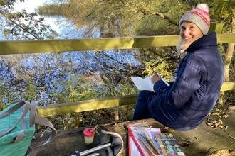 Lady sat on a bench overlooking a body of water, with a pad of paper, paint pallete and various art materials, smiling over her shoulder back at the camera
