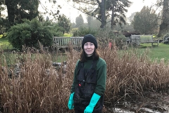 A woman in waders and bright blue rubber gloves stand knee-deep in a pond with a big grin on her face