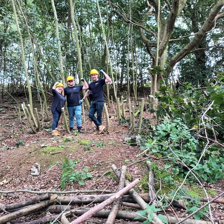 3 members of staff from SUEZ on a Wild Work Day, working in a woodland area, holding their arms up in a muscle pose towards the camera