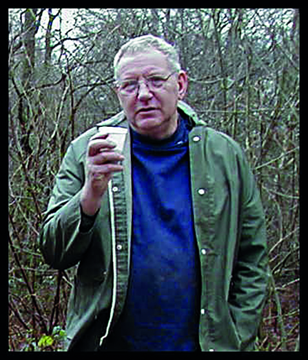 A man wearing a sage green raincoat holds up a cup in a 'cheers' pose against a backdrop of bare winter woody scrub