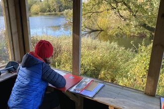 Lady sitting in a hide, looking out to the water with her art materials for journaling