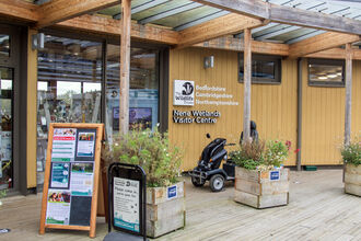 The front door to the Nene Wetlands visitor centre with wooden plant pots outside next to a couple notice boards and a mobility scooter