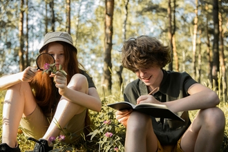 Teenage boy and girl sat under trees identifying flowers with a magnifying glass