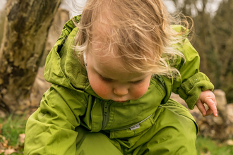 toddler wearing green puddlesuit and blue wellies reaching down to pick up stick