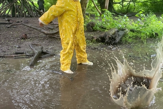A toddler in yellow rain suit throws something into a muddy puddle causing a large splash