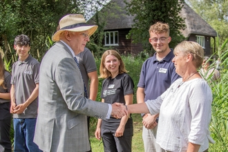 The Duke of Gloucester shakes hands with Louise Rackham, as Wildlife Trust staff and volunteers stand in a line with Rothschild Bungalow in the background.  
