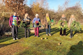 Six people stood in the sunshine holding Christmas wreaths decorated with evergreen foliage and ribbon