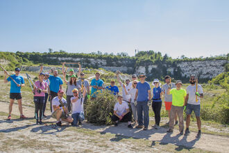Group of people outdoors, smiling while holding various tools including hedge clippers, trowels and axes. They are gathered around a pile of bush cuttings - it is a sunny day. 
