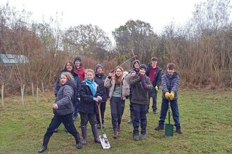 A group of young people dressed up in winter coats and hats smiling at the camera infront of some small trees. Some are holding shovels.