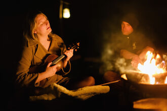 A woman plays a ukelele at a campfire while singing.