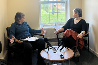Two women sit with microphones on a table in front of them, having a conversation