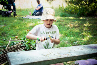 A child in the foreground wearing a floppy cream sunhat using a potato peeler to scrape away the bark of the elder twig on a summers day, whilst sat in the shade. Out of focus behind there is another family with picnic rug and pushchair. A bundle of sticks lays on the ground to one side of the child.
