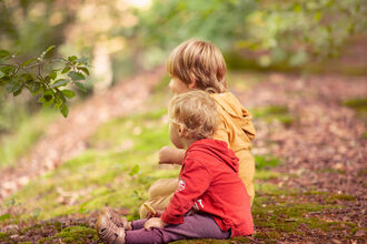 two toddlers sitting on a woodland track facing sideways looking into a woodland, one younger toddler in the foreground, an older one behind. The ground is mossy and leaf strewn.