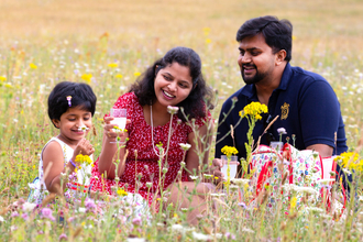 Indian family of mother, father and daughter sit enjoying a picnic in a wildflower meadow. The child is smelling an oxeye daisy. Everyone is smiling