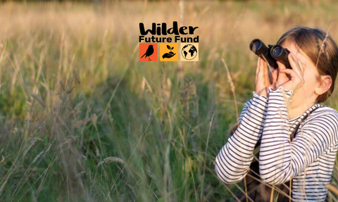 A young girl looks through binoculars on a grassy meadow. The Wilder Future Fund logo is displayed in the middle.