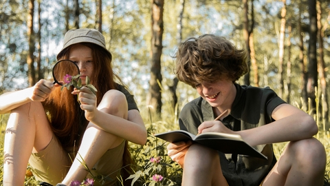 Teenage boy and girl sat under trees identifying flowers with a magnifying glass