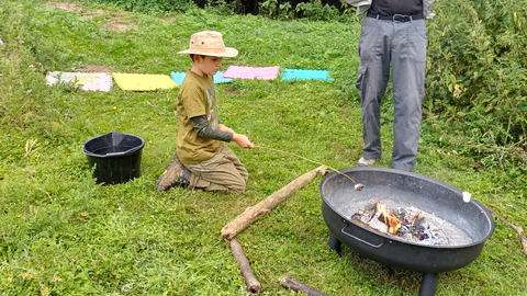 A boy kneels toasting a marshmallow on a stick over a fire pit. An adult stands observing.
