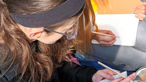 Two youths sat next to each other on a tarpaulin outdoors, their faces hidden, sketching with pencils