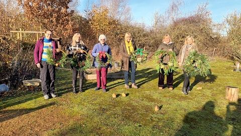 Six people stood in the sunshine holding Christmas wreaths decorated with evergreen foliage and ribbon