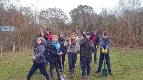 A group of young people dressed up in winter coats and hats smiling at the camera infront of some small trees. Some are holding shovels.