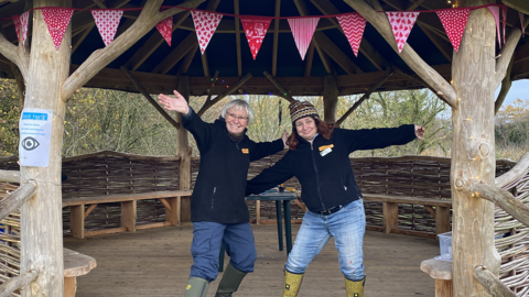 Caroline and Claire stand arms open wide in a wooden shelter decorated with bunting