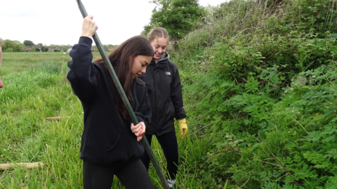 Two girls standing side-by-side in a field, one using a long pole to create a hole in the ground.