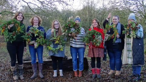 A group of 7 women stood outside showing willow wreaths that they have made from plants and decorations