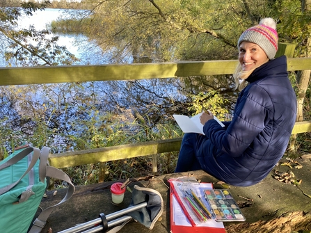 Lady sat on a bench overlooking a body of water, with a pad of paper, paint pallete and various art materials, smiling over her shoulder back at the camera