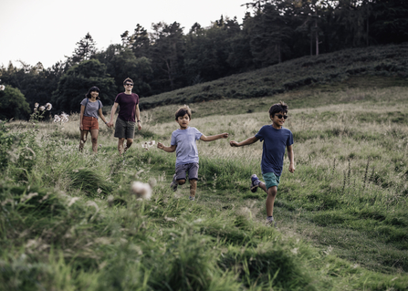 Two children run, smiling, ahead of two adults along a grassy path in a field edged with trees