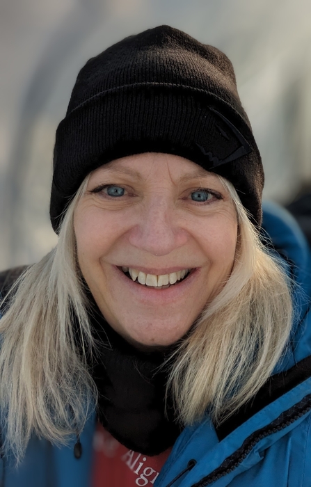 A woman with mid-length blonde hair, wearing a black beanie hat, smiles broadly at the camera