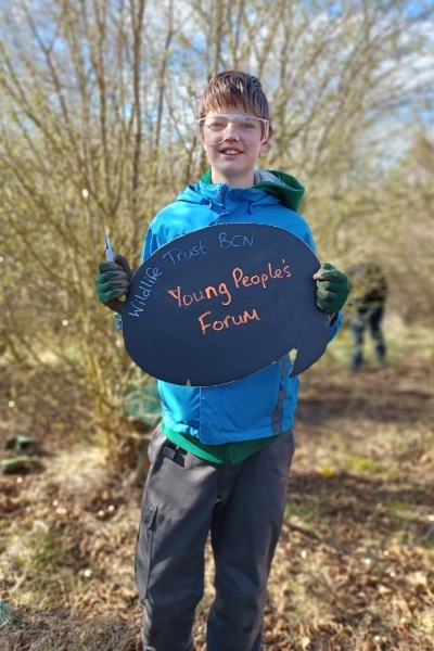 Callum, YPF member, at Cambourne Community Garden on a sunny day, holding up a blackboard sign with "Wildlife Trust BCN Young People's Forum" written on it