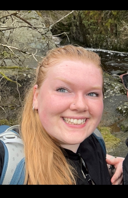 A woman wearing a backpack with long orange hair tied into a ponytail smiles at the camera as she takes a selfie