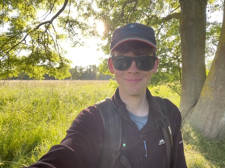 A young man wearing sunglasses and a cap smiles at the camera as he stands outside on a bright, sunny day