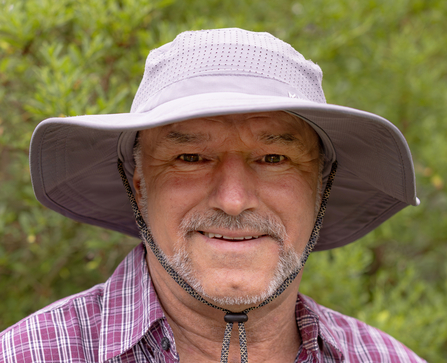 A man with a short, grey beard, wearing a wide-brimmed sun hat, smiles at the camera against a background of green foliage