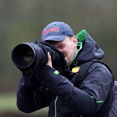 A man in a waterproof coat and a 'PNG' cap looks through the viewfinder of a camera with a large lens, smiling