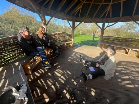 Two people sit on a bench, with a third on the floor in front of them, in an outdoor pavilion