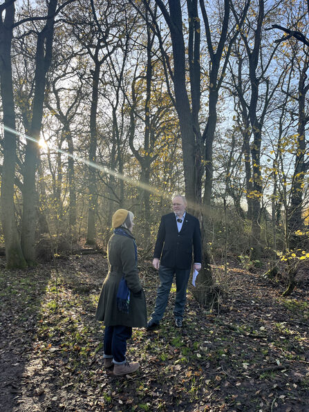 A man and a woman stand chatting in a wintry woodland, surrounded by large elm trees