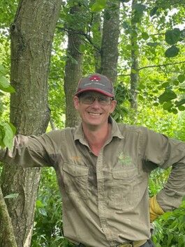 A man smiles while he leans against a tree in a wooded area