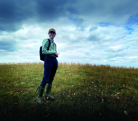 A young man stands in grassland, a dramatic clouded sky behind him