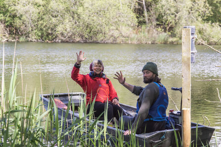 A woman in a red coat and a man in a green hat wave at the camera from a punt in the reeds as they head out onto the water