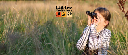 A young girl looks through binoculars on a grassy meadow. The Wilder Future Fund logo is displayed in the middle.