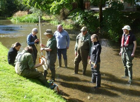 A group of 8 people in waders and wellington boots standing in a shallow river on a training course
