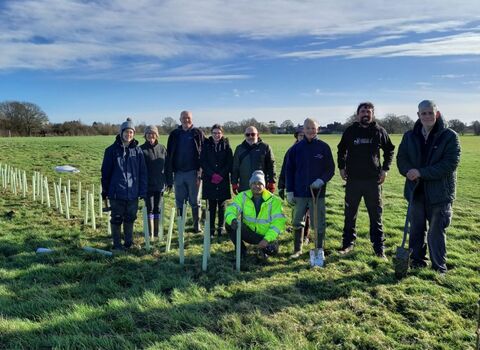 A goroup of people standing in a field alongside a row of planted trees, smiling at the camera