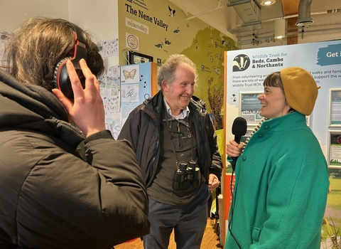 A woman in a yellow hat smiles as she chats to a man with binoculars round his neck, another woman with headphones in the foreground