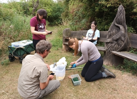 Three work experience students help with surveying newt traps with Henry. One takes photos on a phone, one sits on the bench recording on a clipboard and one kneels to open to traps