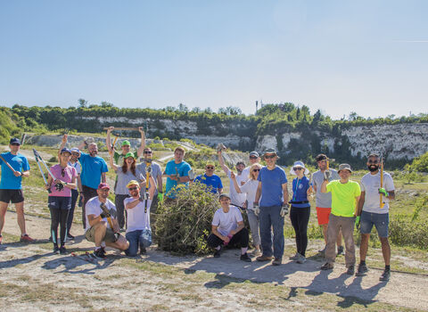 Group of people outdoors, smiling while holding various tools including hedge clippers, trowels and axes. They are gathered around a pile of bush cuttings - it is a sunny day. 