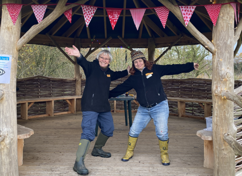 Caroline and Claire stand arms open wide in a wooden shelter decorated with bunting