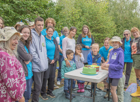 A group of people of various ages gather around a celebration cake and smile at the camera